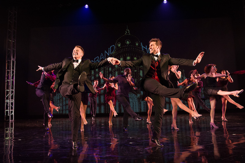 Two male dancers in bow ties, vests and tails link arms and kick enthusiastically with the chorus members behind them